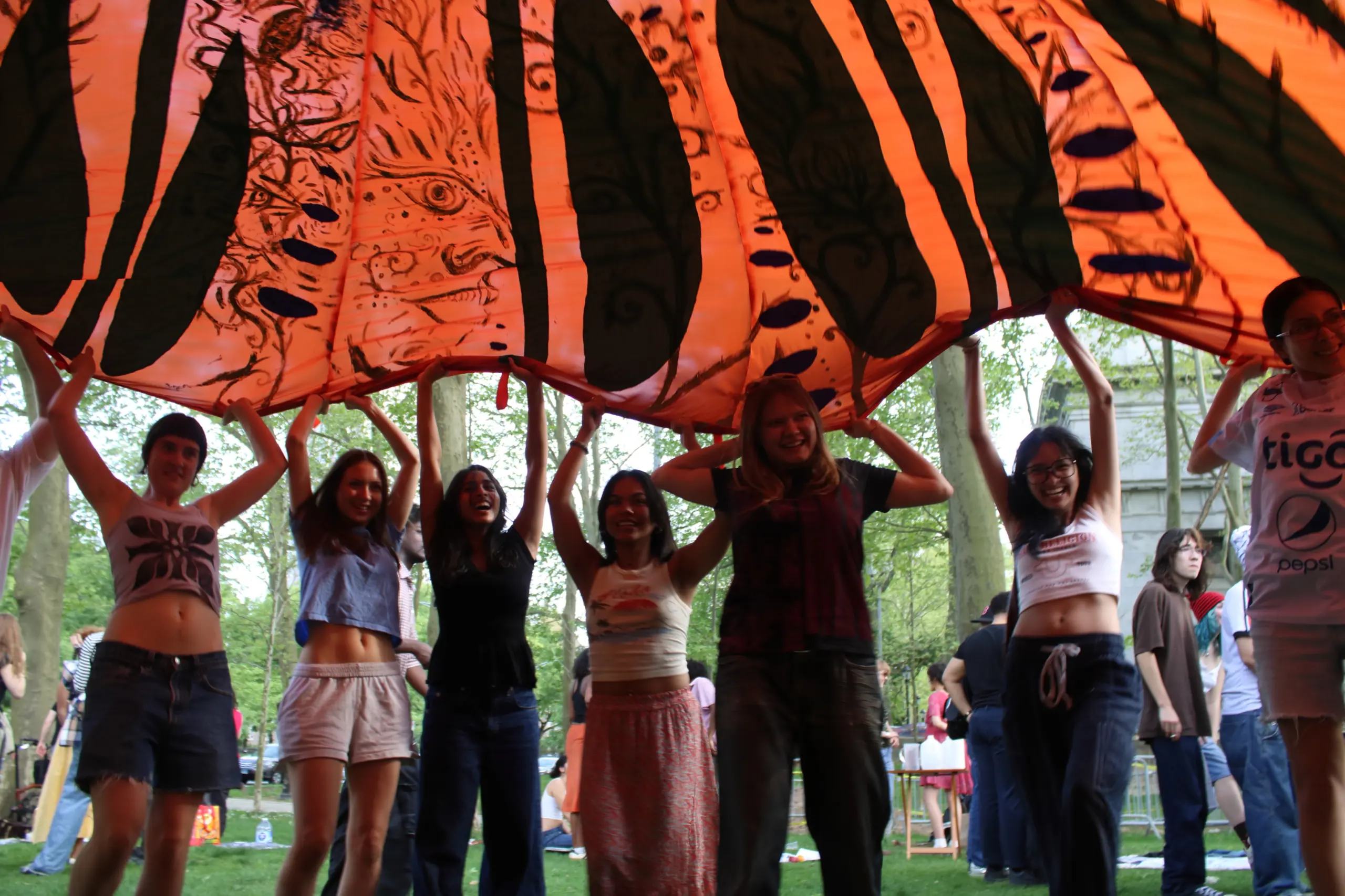 Young people outdoors in a park, lifting a very large piece of fabric up over their heads like a parachute 