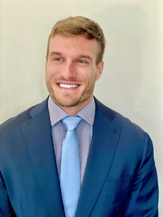 Blonde man smiling into camera on tan background. He is wearing a dark blue suit jacket over a light shirt with a light blue tie.