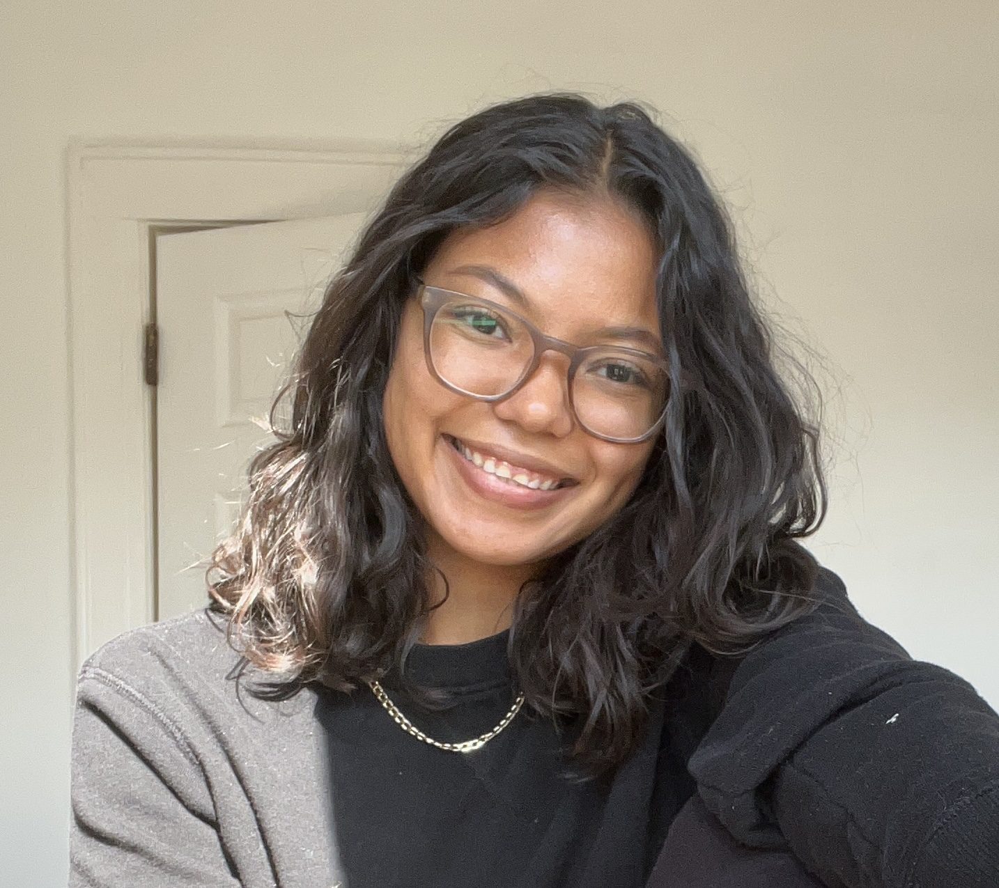 A smiling person with wavy sholder length brown hair, glasses, a black sweatshirt and sliver chain necklace, against a cream colored door.