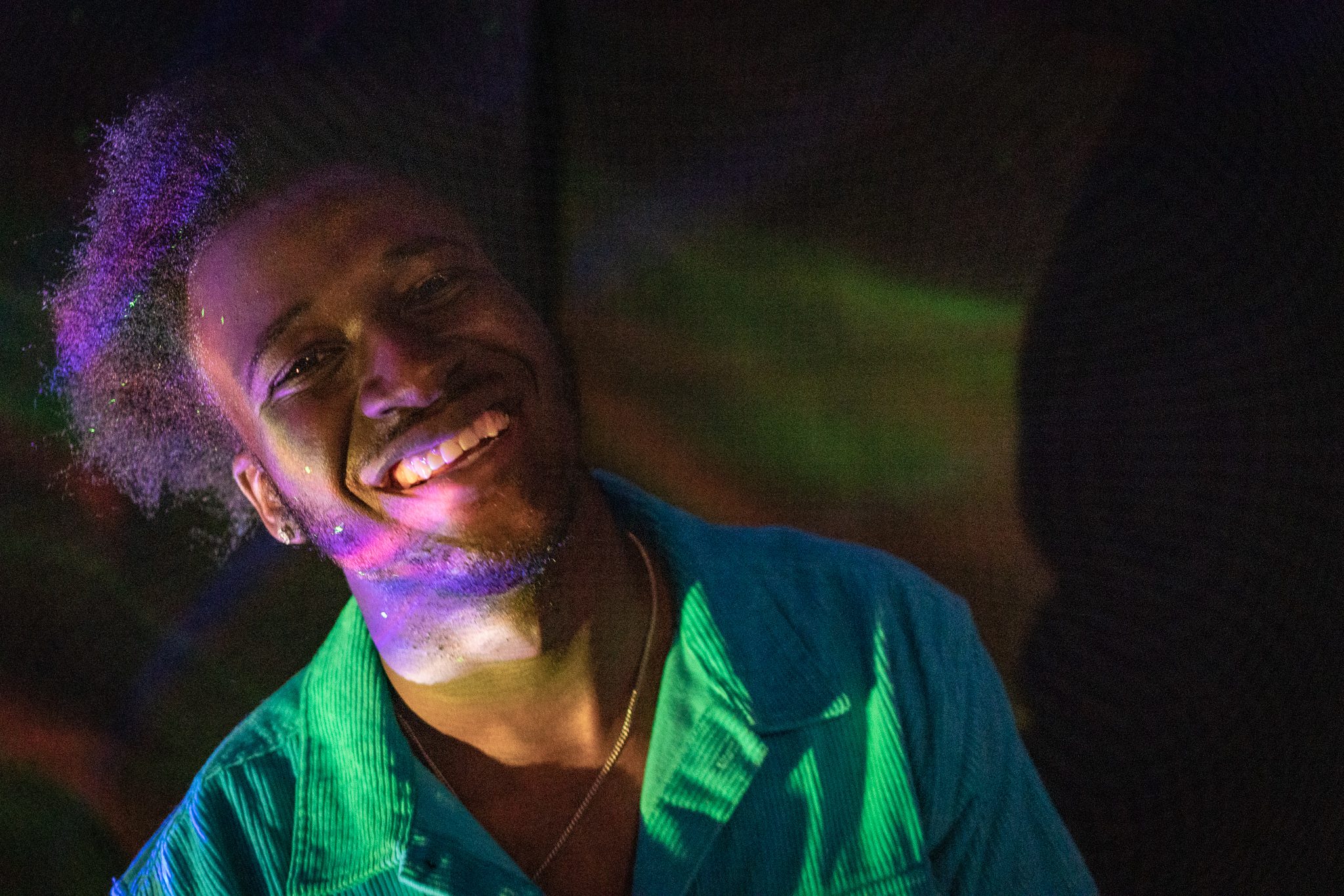 A smiling person leaning to the left, wearing a green button down shirt and gold chain, bathed in rainbow light with and iridescent black background.