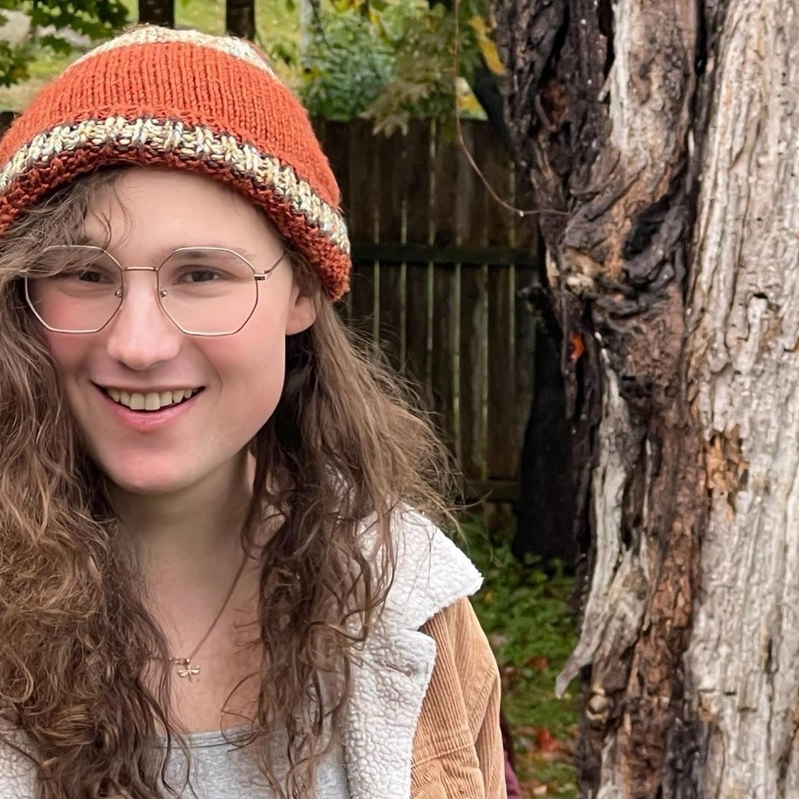 Portrait of a person with long wavy hair and glasses wear a hat and jacket standing outside beside a tree.