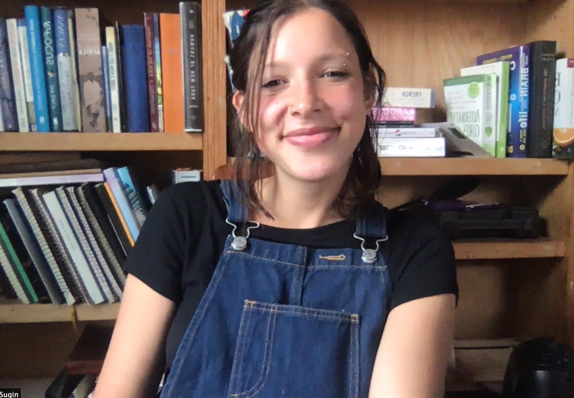 Aperson in a black teeshirt and dark blue jean overalls with short brown hair pulled back sitting in front of a bookshelf.