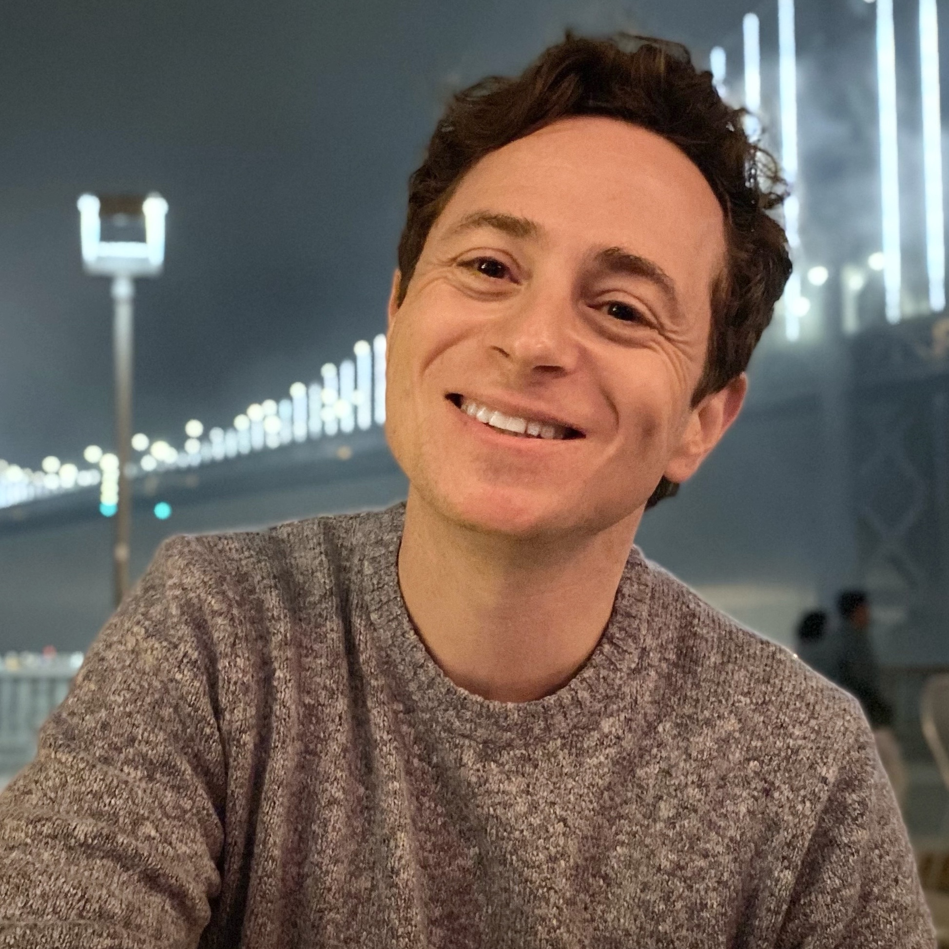 A person smiling at the camera with short wavy brown hair and a heather grey sweater sitting in front of a bridge with bright lights at night.