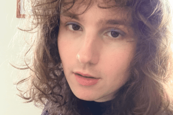A person with curly brown hair wearing a black turtle neck and blie eyeshadow.