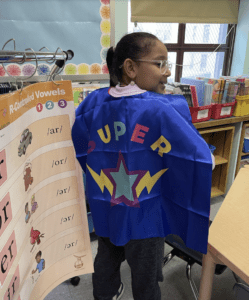 Second grade student in a Bronx classroom wearing a blue cape with SUPER written in in bright colors with lightning bolts