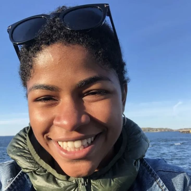 A close headshot of Dominique smiling with water behind them.