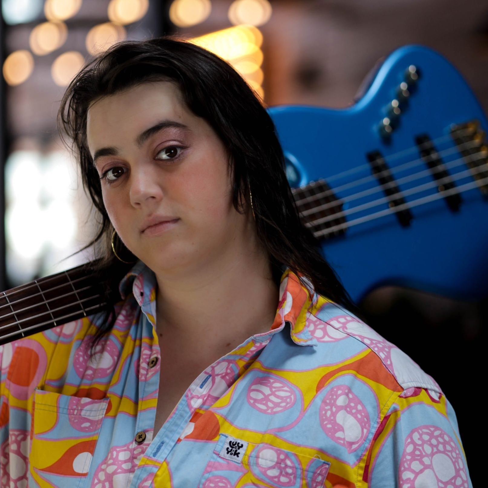 A photo of Lulu in a multicolored shirt with a blue guitar over their shoulder.