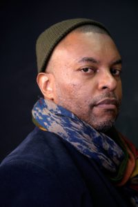 Portrait of artist in a beanie and scarf shown in three quarters profile again black background.