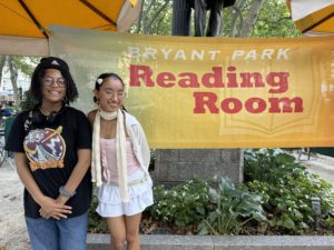 Two high school students stand before a banner at NYC's Bryant Park Reading Room during a bright summer day.