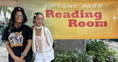 Two high school students stand before a banner at NYC's Bryant Park Reading Room during a bright summer day.