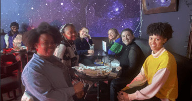 Women artists gathered at an Art Salon in room with walls painted with the sky at night with stars.