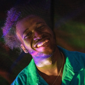 A smiling person leaning to the left, wearing a green button down shirt and gold chain, bathed in rainbow light with and iridescent black background.