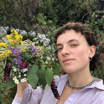 Woman with in lilac shirt with a bouquet of wildflowers near her face and green garden and trews in the background