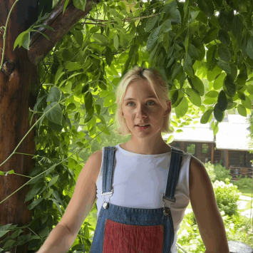 Woman in overalls in front of trees looking at camera