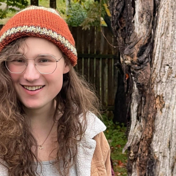 Portrait of a person with long wavy hair and glasses wear a hat and jacket standing outside beside a tree.