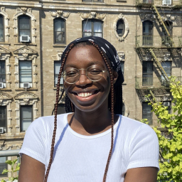 Black woman smiling in bright sun wearing white tshirt with NYC apartment building as the background