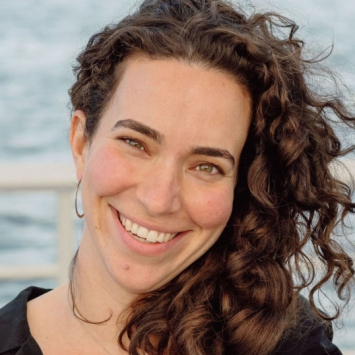 Close up of the smiling face of a person with curly brown hair, black shirt and sliver hoop earrings