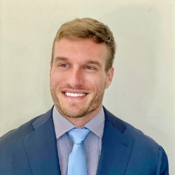 Blonde man smiling into camera on tan background. He is wearing a dark blue suit jacket over a light shirt with a light blue tie.